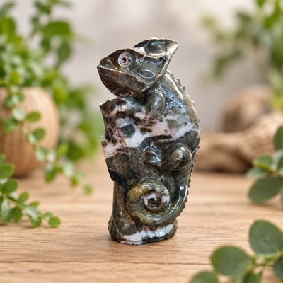 Side angle of a natural jade chameleon carving on a light wooden table with greenery.