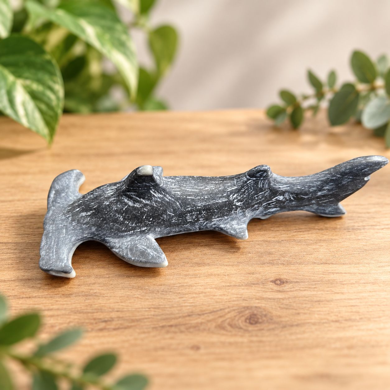 Taiji stone hammerhead shark carving lying on a light wooden table with greenery.
