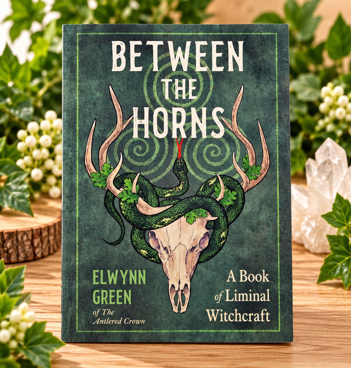 Between the Horns: A Book of Liminal Witchcraft by Elwynn Green displayed on a light wooden table with greenery.