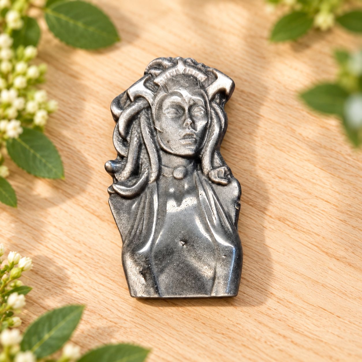 Silver Sheen Obsidian Medusa carving displayed flat on a light wooden table with greenery