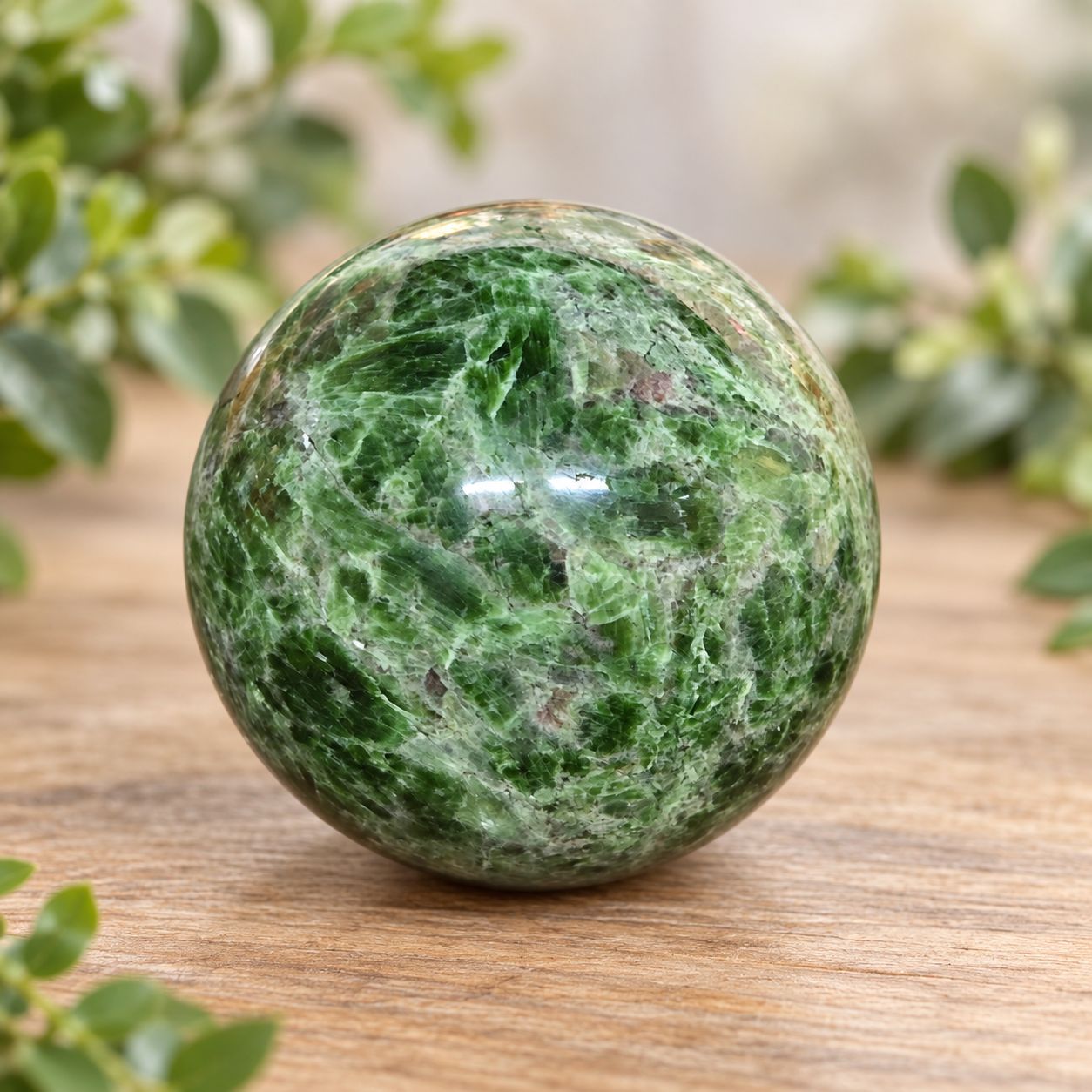 Polished green Diopside sphere displayed on a light wooden table with greenery