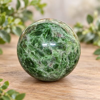 Polished green Diopside sphere displayed on a light wooden table with greenery