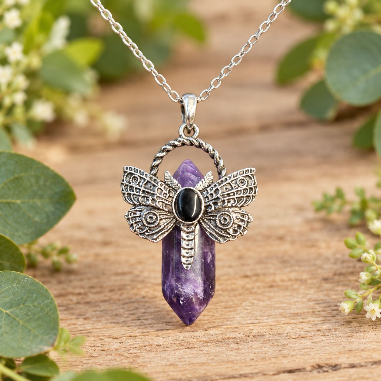 Amethyst dragonfly point necklace with silver-tone pendant and stainless steel chain on light wood with greenery.