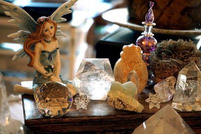Fairy figurine with blue wings sitting on a wooden surface surrounded by clear quartz crystals, an owl carving, a small nest,