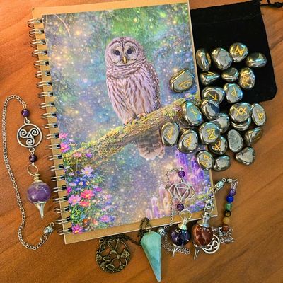 Owl‑illustrated notebook with rune stones, crystals, and pendulums arranged around it