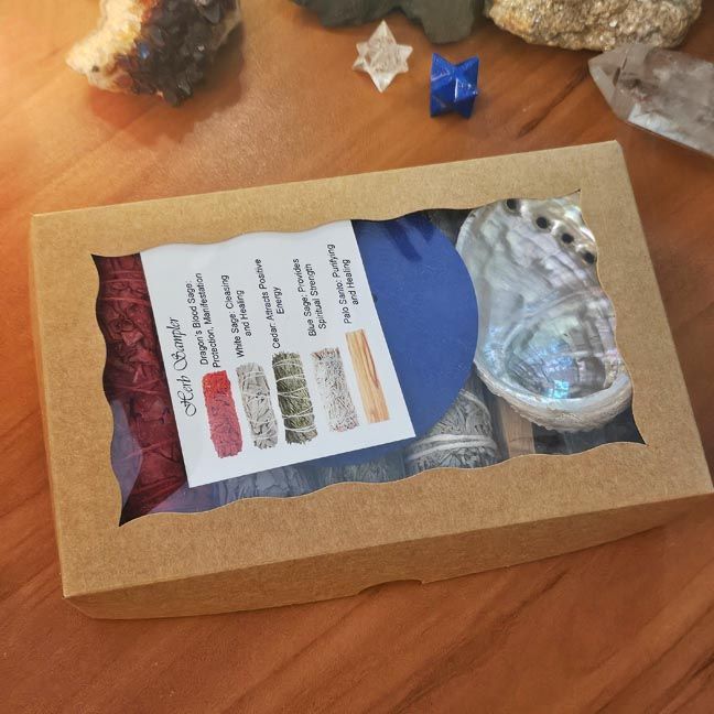 Smudging sampler box with sage bundles, palo santo, selenite wand, and abalone shell displayed on a wooden surface.