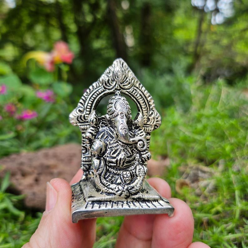 Small silver Ganesh statue held between fingers outdoors, with green foliage and flowers softly blurred in the background.