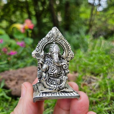Small silver Ganesh statue held between fingers outdoors, with green foliage and flowers softly blurred in the background.