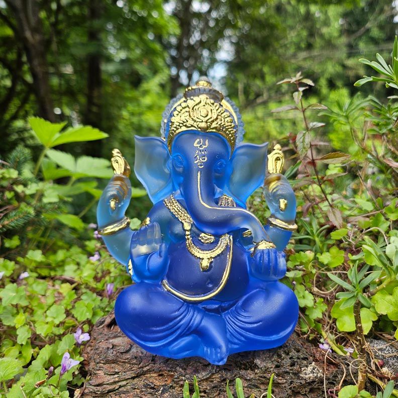 Blue Ganesh statue with gold accents seated on a wooden surface outdoors, surrounded by green plants.