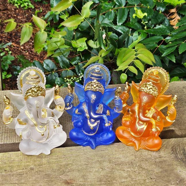 Three Ganesh figurines in white, blue, and orange with gold accents displayed outdoors on a wooden surface with green leaves