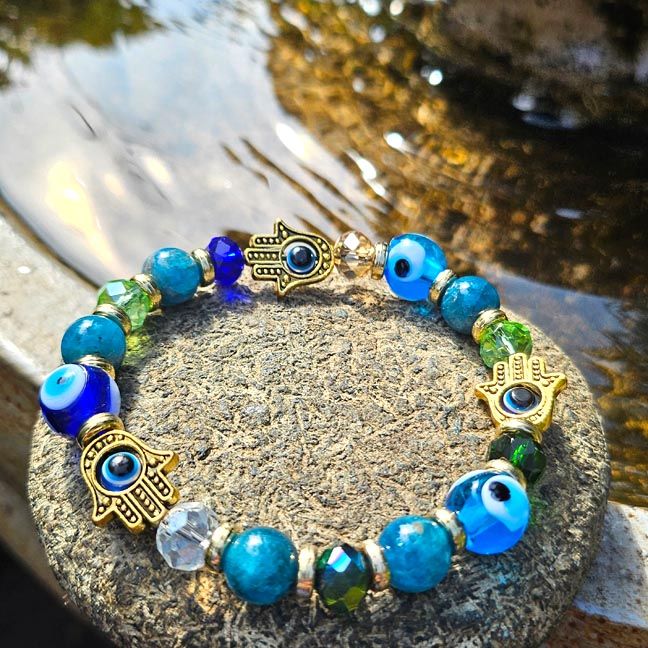 Bracelet with blue, green, and clear beads and small gold Hamsa charms, displayed on a stone near water in sunlight.