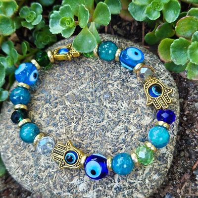 Bracelet with blue, turquoise, green, and gold beads and small gold Hamsa charms, displayed on a stone with succulents