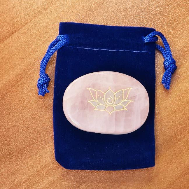 Rose quartz palm stone with a gold Lotus engraving placed on a blue velvet pouch on a wooden surface.