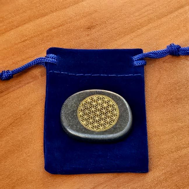 Hematite palm stone with a gold Flower of Life engraving placed on a blue velvet pouch on a wooden surface.