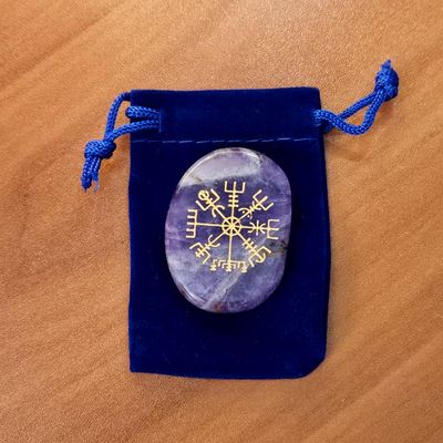 Amethyst palm stone with a gold Vegvisir engraving placed on a blue velvet pouch on a wooden surface.