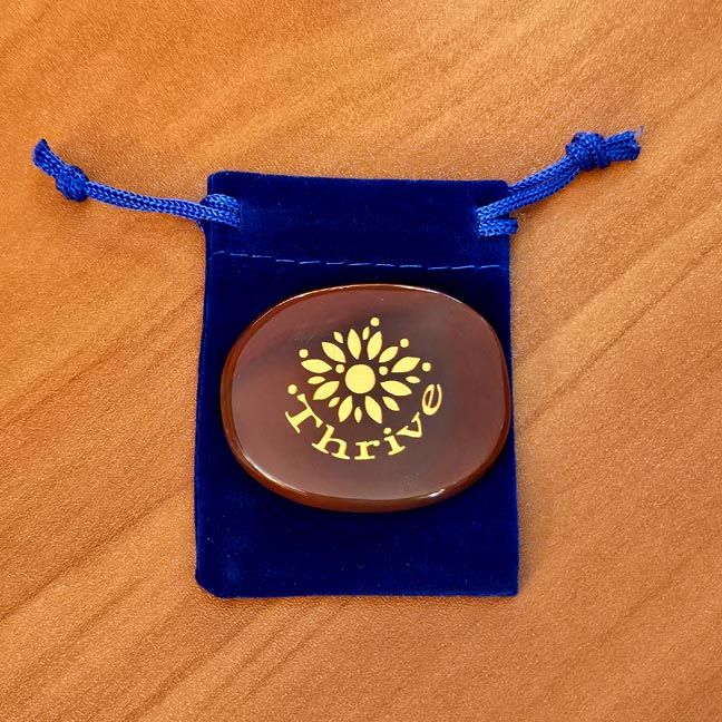 Carnelian palm stone with a gold flower and the word Thrive engraving placed on a blue velvet pouch on a wooden surface.