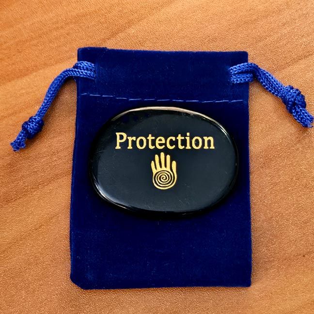 Obsidian palm stone with the word ‘Protection’ and a hand symbol engraving placed on a blue velvet pouch on a wooden surface.