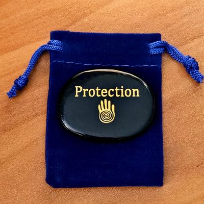 Obsidian palm stone with the word ‘Protection’ and a hand symbol engraving placed on a blue velvet pouch on a wooden surface.