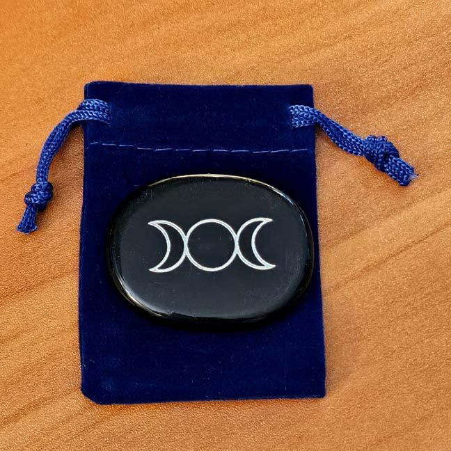 Obsidian palm stone with Triple Moon engraving placed on a blue velvet pouch on a wooden surface.