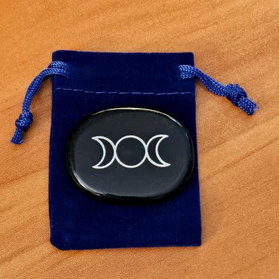 Obsidian palm stone with Triple Moon engraving placed on a blue velvet pouch on a wooden surface.