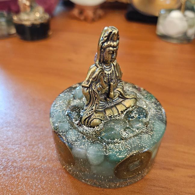 Small Kuan Yin brass figure on a round resin base with green stones and gold glitter, displayed on a wooden surface indoor