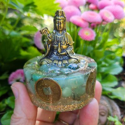 Small brass Kuan Yin on a round resin base with green stones, gold glitter, and coins, held outdoors near pink flowers.