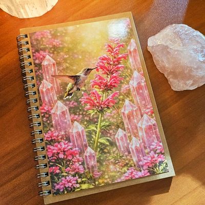 Spiral‑bound journal with a hummingbird hovering near pink flowers surrounded by glowing pink crystals in a dreamy garden