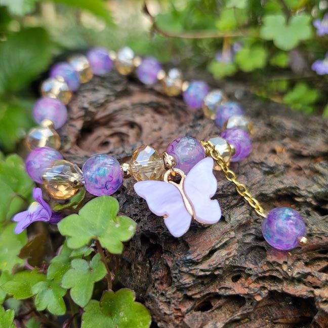 Bracelet with purple beads, faceted gold beads, and a lavender butterfly charm on a gold chain, on a brown stone with plants