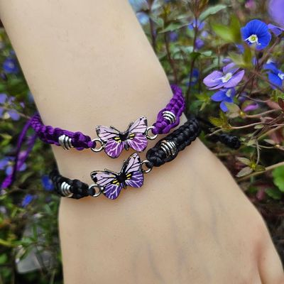 Wrist wearing two woven bracelets with purple butterfly charms, with small purple and blue flowers and green foliage.