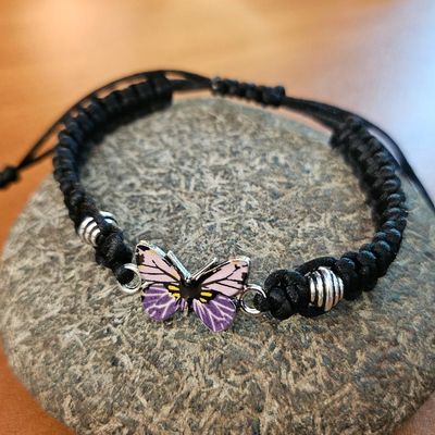Black braided cord bracelet with a purple butterfly charm and small metallic beads, displayed on a smooth stone.