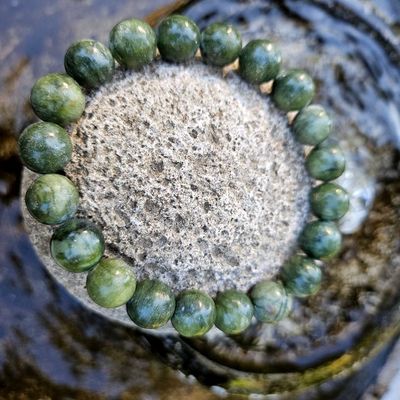 Green jade bead bracelet resting on a textured gray rock beside water in an outdoor setting.