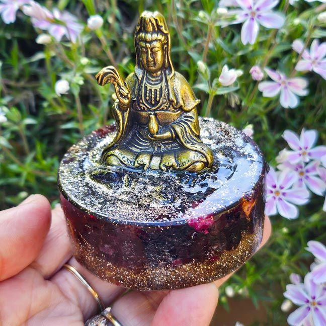 Small brass seated Kuan Yin on a round resin base with red tones and glitter, held in a hand outdoors near green foliage.