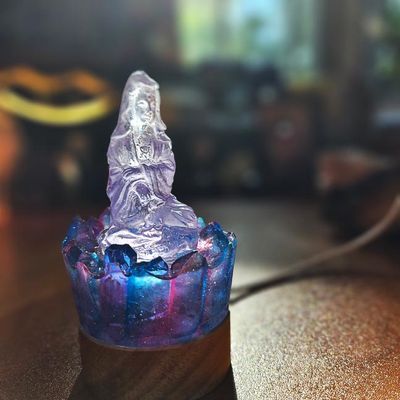 Translucent seated Kuan Yin on a blue‑purple crystal base with a wooden stand, softly glowing in warm light