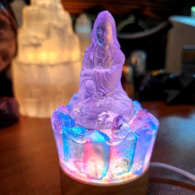 Translucent seated Kuan Yin glowing with shifting blue, pink, and purple light atop a crystal‑like illuminated base