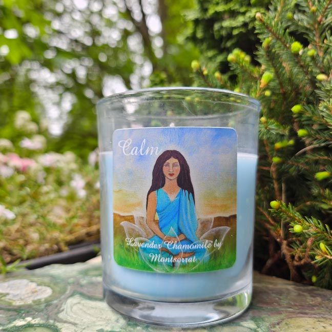 Blue candle labeled “Calm – Lavender Chamomile by Montserrat,” placed outdoors on a green stone with greenery