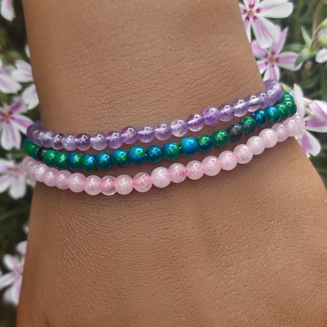 Three thin gemstone bracelets on a wrist, in rose quartz, blue‑green chrysocolla, and amethyst with soft floral background