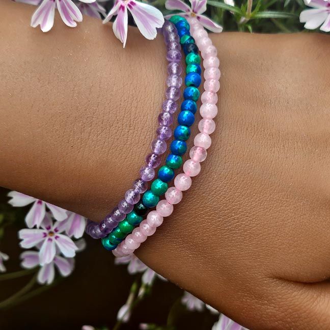 Three thin gemstone bracelets on a wrist, in rose quartz, blue‑green chrysocolla, and amethyst with soft floral background