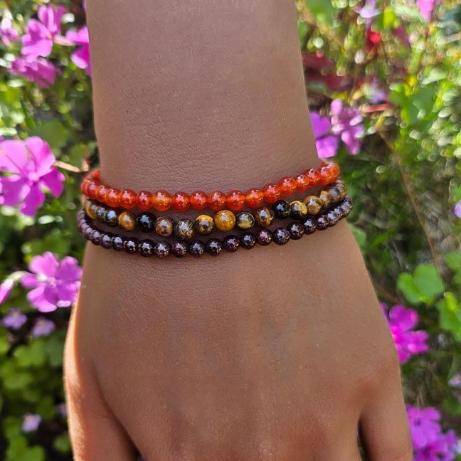 Three gemstone bracelets — Garnet, Carnelian, and Tiger Eye — glowing in sunlight against garden flowers.