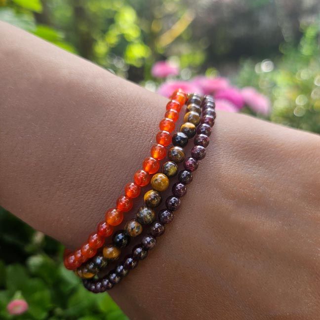 A wrist adorned with three gemstone bracelets: deep red Garnet, fiery orange Carnelian, and golden Tiger Eye 