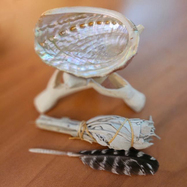 Small abalone shell on a wooden tripod beside a bundled white sage wand and a black‑and‑white feather on a wooden surface.