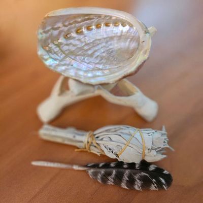 Small abalone shell on a wooden tripod beside a bundled white sage wand and a black‑and‑white feather on a wooden surface.