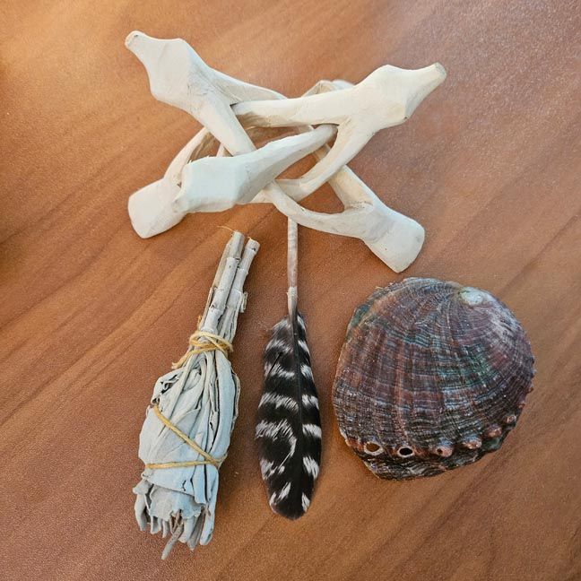 Small abalone shell,a wooden tripod beside a bundled white sage wand and a black‑and‑white feather on a wooden surface.