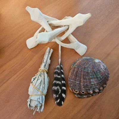 Small abalone shell,a wooden tripod beside a bundled white sage wand and a black‑and‑white feather on a wooden surface.