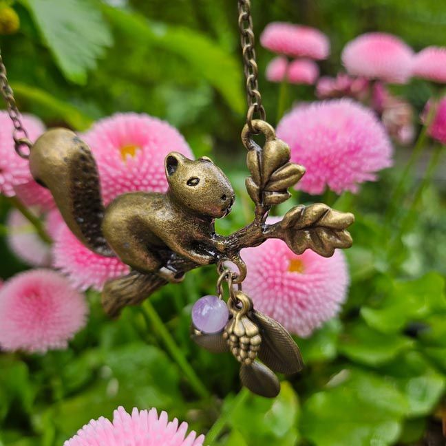 Bronze squirrel necklace on an oak branch with leaves, pine cone, and lilac amethyst bead among pink flowers
