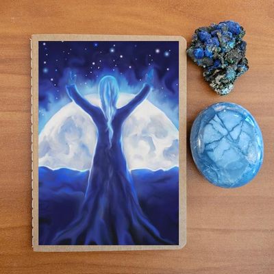 Notebook with an illustration of a robed figure standing before a glowing moon, on a wooden surface with crystals