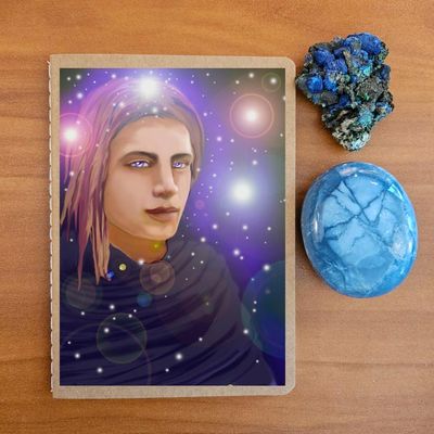 Notebook with an illustration of a long‑haired figure against a cosmic background, on a wooden surface with crystals