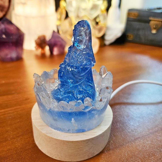 Pale blue resin Kuan Yin figurine surrounded by clear quartz points on a wooden LED base.