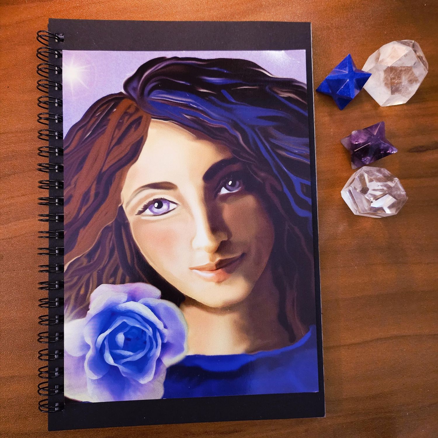 Spiral‑bound art journal with a painted woman holding a blue rose, shown beside crystals on a wooden surface.