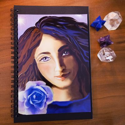 Spiral‑bound art journal with a painted woman holding a blue rose, shown beside crystals on a wooden surface.