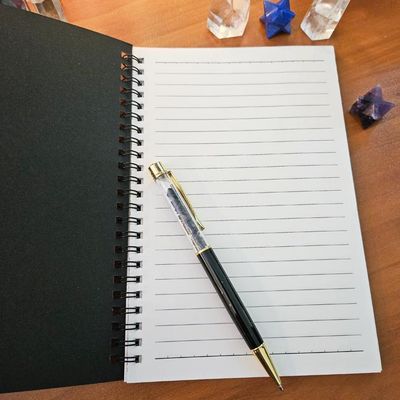 Open spiral‑bound lined journal with a black cover and pen resting diagonally, surrounded by crystals on a wooden surface.